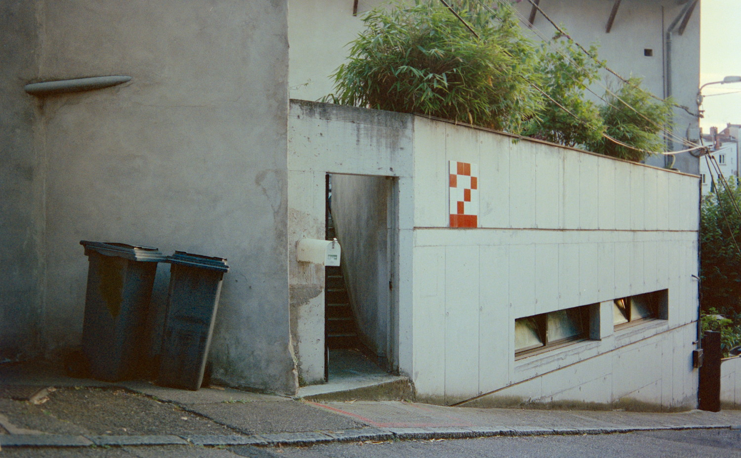 Wandering through the narrow street of a residential neighbourhood, I passed a corner and immediatly saw there was a shot to take here. You got this very brutalist house facade in that sloppy street, large flat concrete surface with a massive red "2" sign made out of tiles. Some bushes appearing over the wall. The door was open revealing some stairs going higher up. I absolutely loved the place right away and I'm very happy to have it captured.