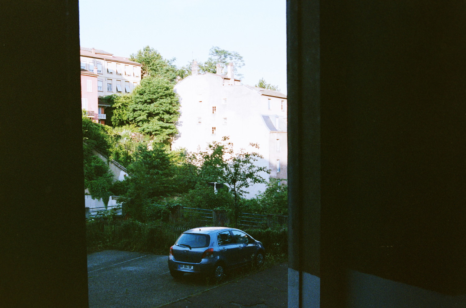 You go down the stair and get greeted by an incredible sunny view. The sun is blasting that building so strong we don't even distinguish it from the sky ! If it was not for the bushy vegetation all around, indicating its silouhette. Vegetation which is actually so lushy we could fear that the car parked near would be quickly covered by it. The place really doesn't feel like a city, small messy and sloping building dispersed through vegation, it feels confy.