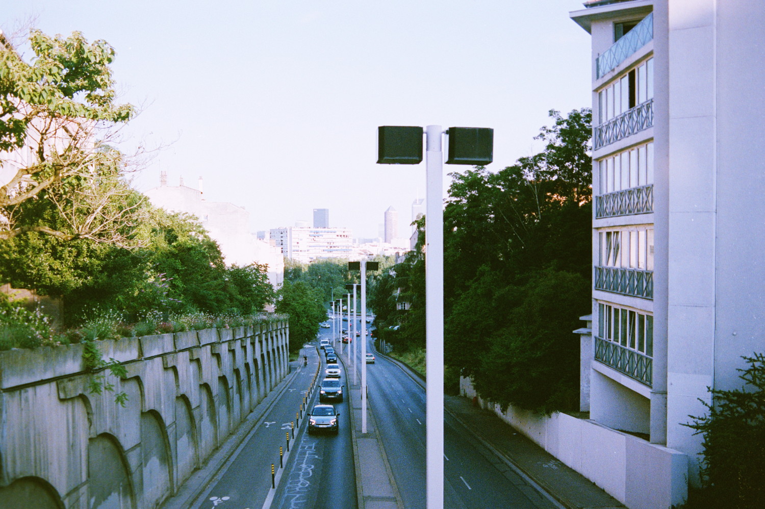 End of the day on the city, people are riding back from work to their home, leading quite some traffic on this popular uphill. On a bridge above it, I capture that line of car. But its no borring road, there is a lot vegetations all around it catching some of the last warmth sun beams. Barely noticeable in the background, we can see the taller building of the financial district that nealy merge with the clear blue sky.