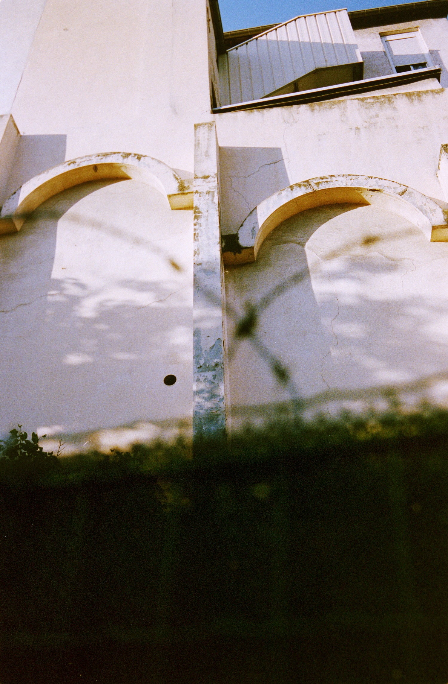 Some funky architecture on the side of a window-less building. Two archs whose seems to not be that recent, catching some of an end-of-day sun. Also noticeable is that foregound, highly out-of-focus but which outline what seems to be a barbed wire fence. There is not much noticeable element in that shot but colors are all over it, with avery pastel palette going from a golden yellow to a washed purple and some very dark greens.