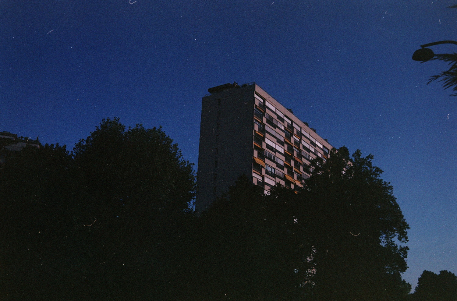 Hidden behind the trees, massive block of concrete. I'm told people lives there, lost in the emptiness of the sky, taking turns for who gets to have the sun. Until it fades away, and all share the noisy night.
