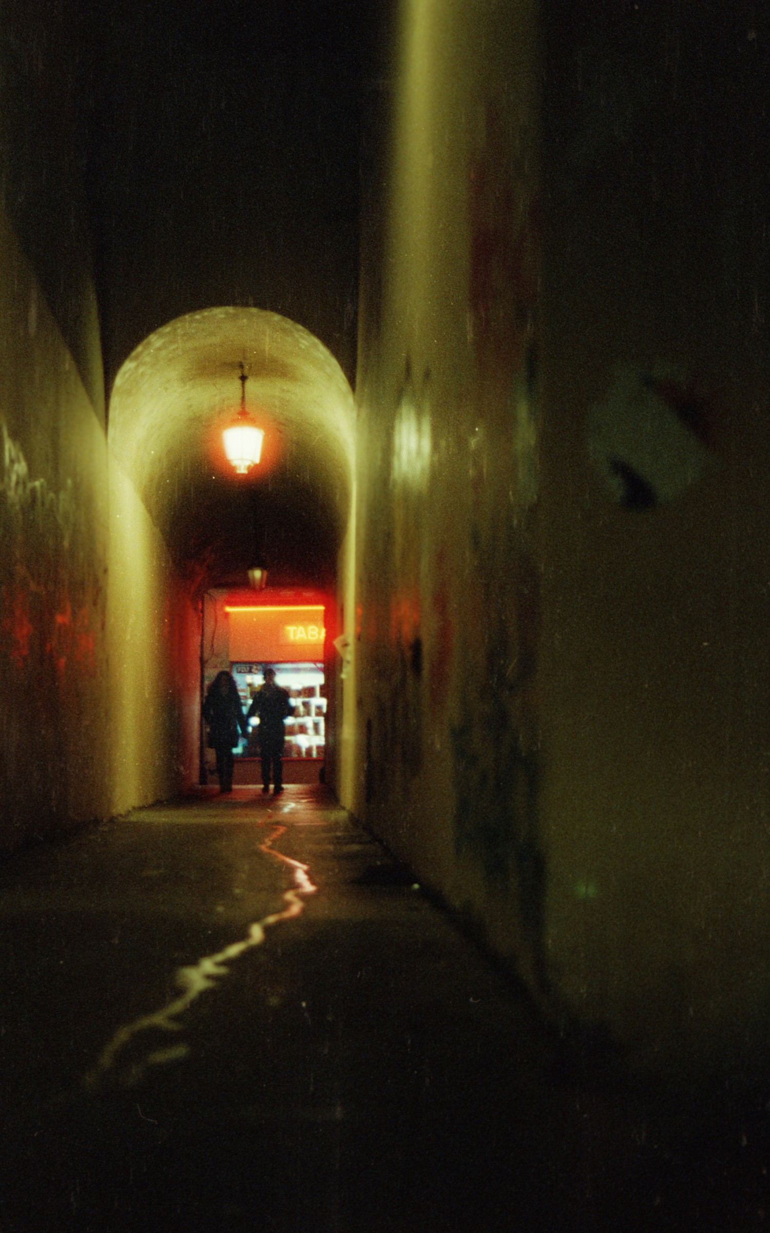 A narrow passage stuck between building. You think it could looks creepy at this time of the night, but its not that dark. A vintage lamp hung at the ceilling is shining strong as it can and reveals the silhouette of a couple holding hands, nicely silhouetted by the bright store-front of a "Tabac" in the background. Not that dark, lovely actually !